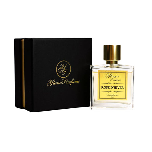 La imagen muestra el perfume "Rose D’Hiver" de Ylusia Parfums presentado junto a su lujosa caja. El frasco, fabricado en vidrio transparente, revela un líquido dorado que resalta su elegancia. La etiqueta dorada en la parte frontal incluye el nombre del perfume y detalles en tipografía sofisticada, reforzando el carácter exclusivo de la fragancia. El tapón metálico dorado completa el diseño con un aire de distinción. La caja negra, con detalles dorados y el logotipo de Ylusia Parfums, refleja el lujo y la
